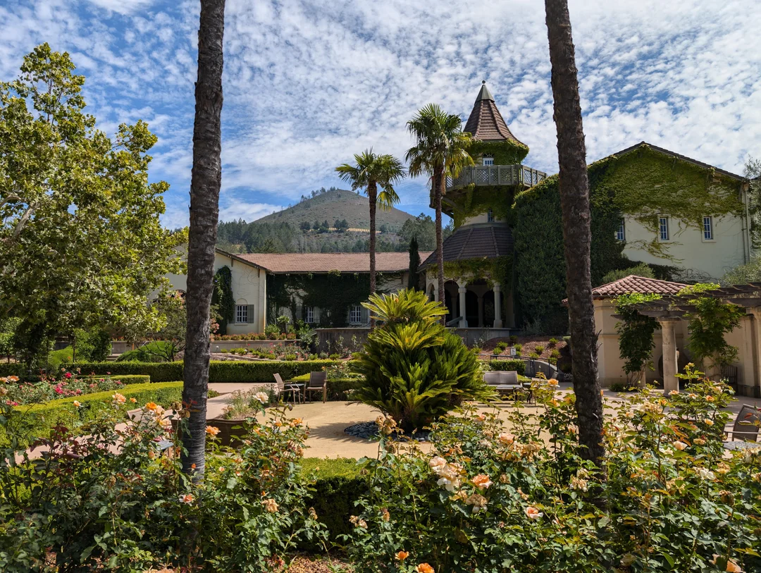 A beautiful garden with blooming flowers, manicured hedges, and tall palm trees sits in front of a large, ivy-covered building with a turret, set against a backdrop of hills and a blue sky with scattered clouds.