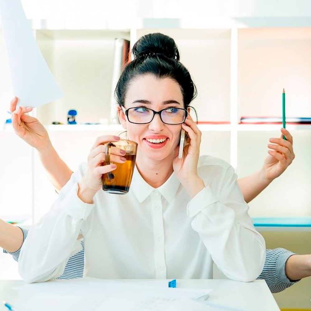 A woman in glasses smiles while talking on the phone and holding a drink, with extra arms appearing to multitask by holding papers, a pen, and a phone in a bright office setting.