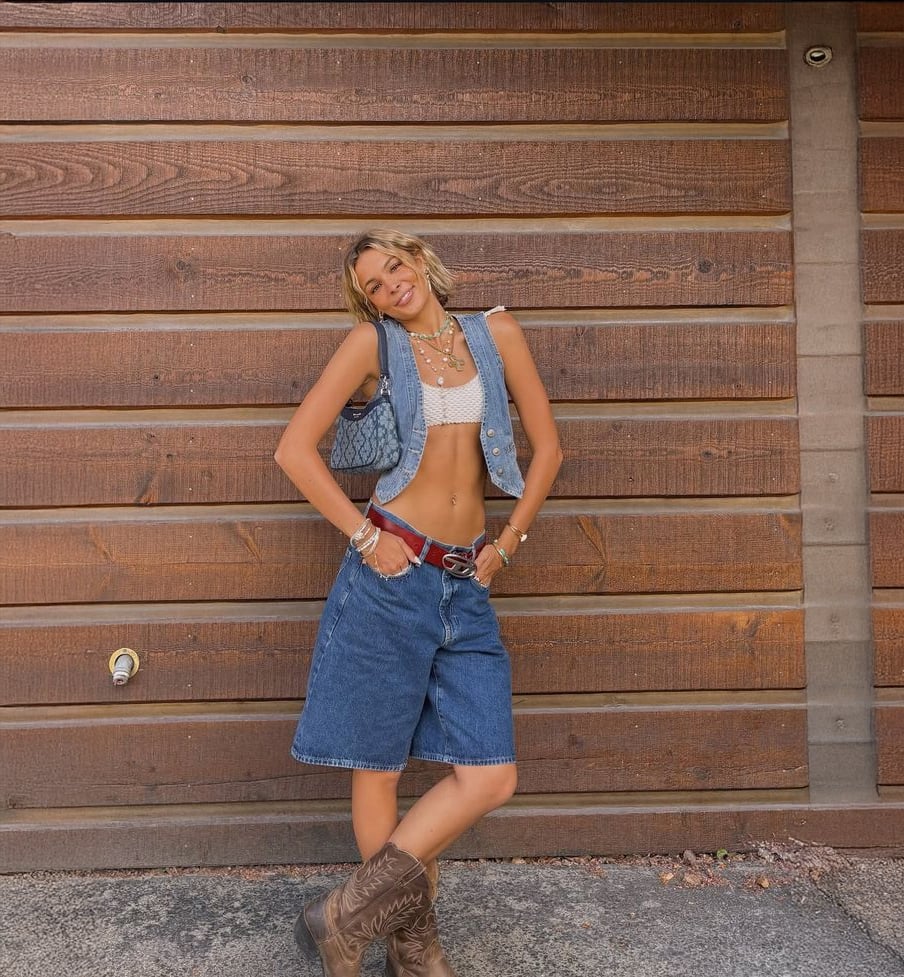 A smiling person stands in front of a wooden wall, wearing a denim vest, white crop top, long denim shorts, and cowboy boots. They accessorize with layered necklaces, a belt, and a shoulder bag.