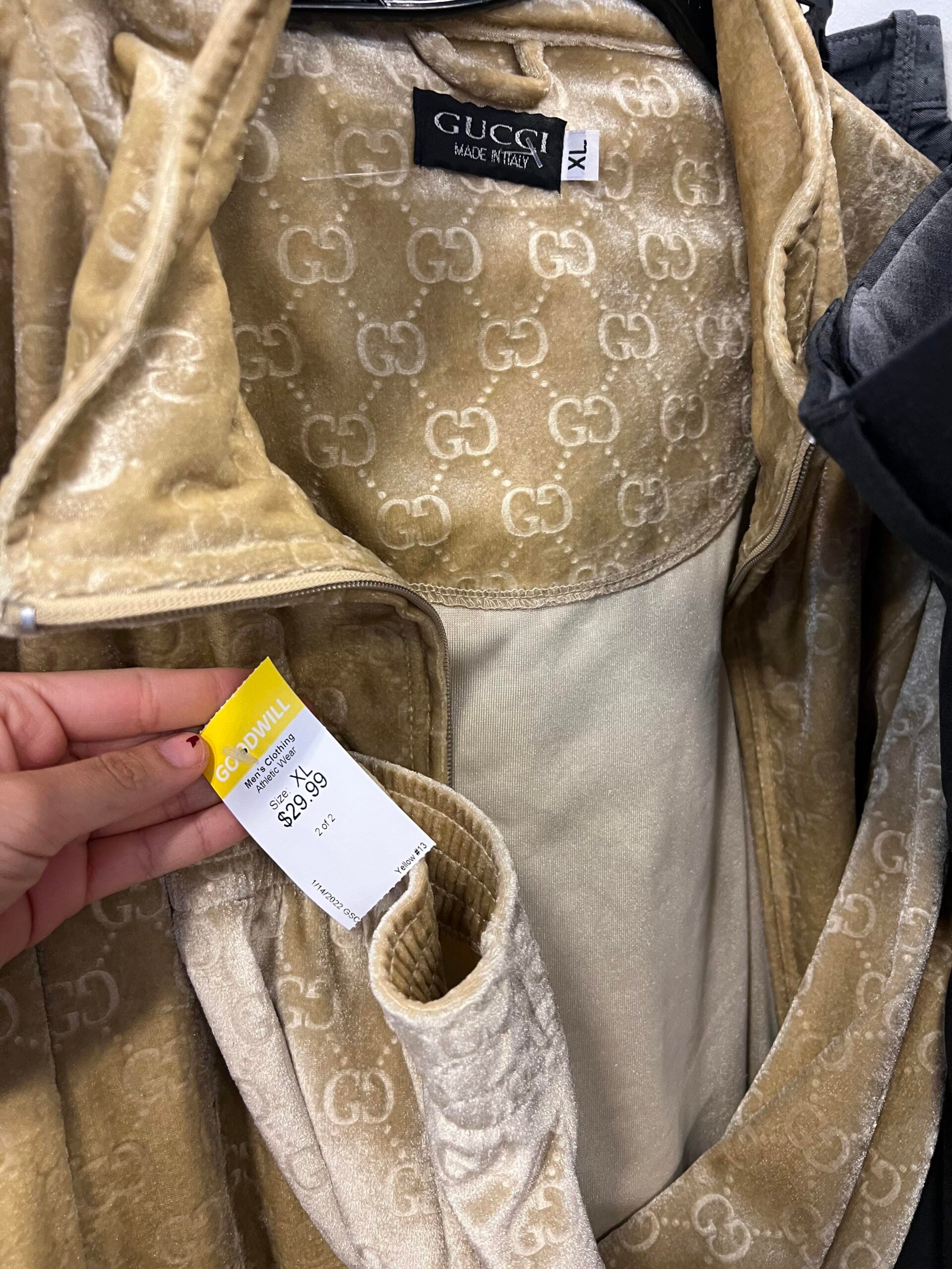A tan velvet Gucci jacket with a repeated "GG" logo pattern hangs on a rack. A hand holds a Goodwill price tag showing $42.99. The inside label reads "GUCCI Made in Italy XL.