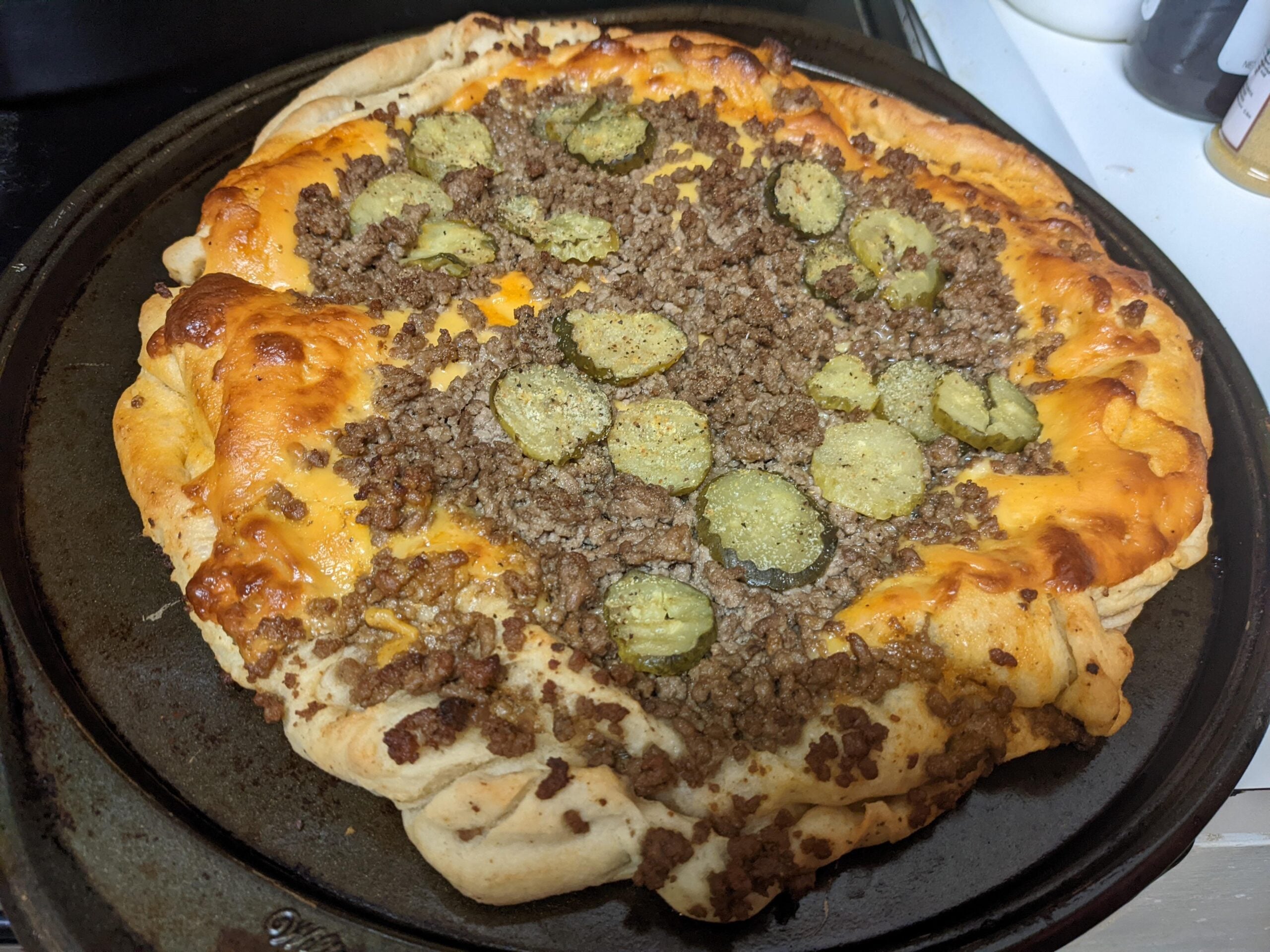 A homemade cheeseburger pizza with a thick crust, topped with ground beef, melted cheddar cheese, and sliced pickles, sitting on a round baking tray.