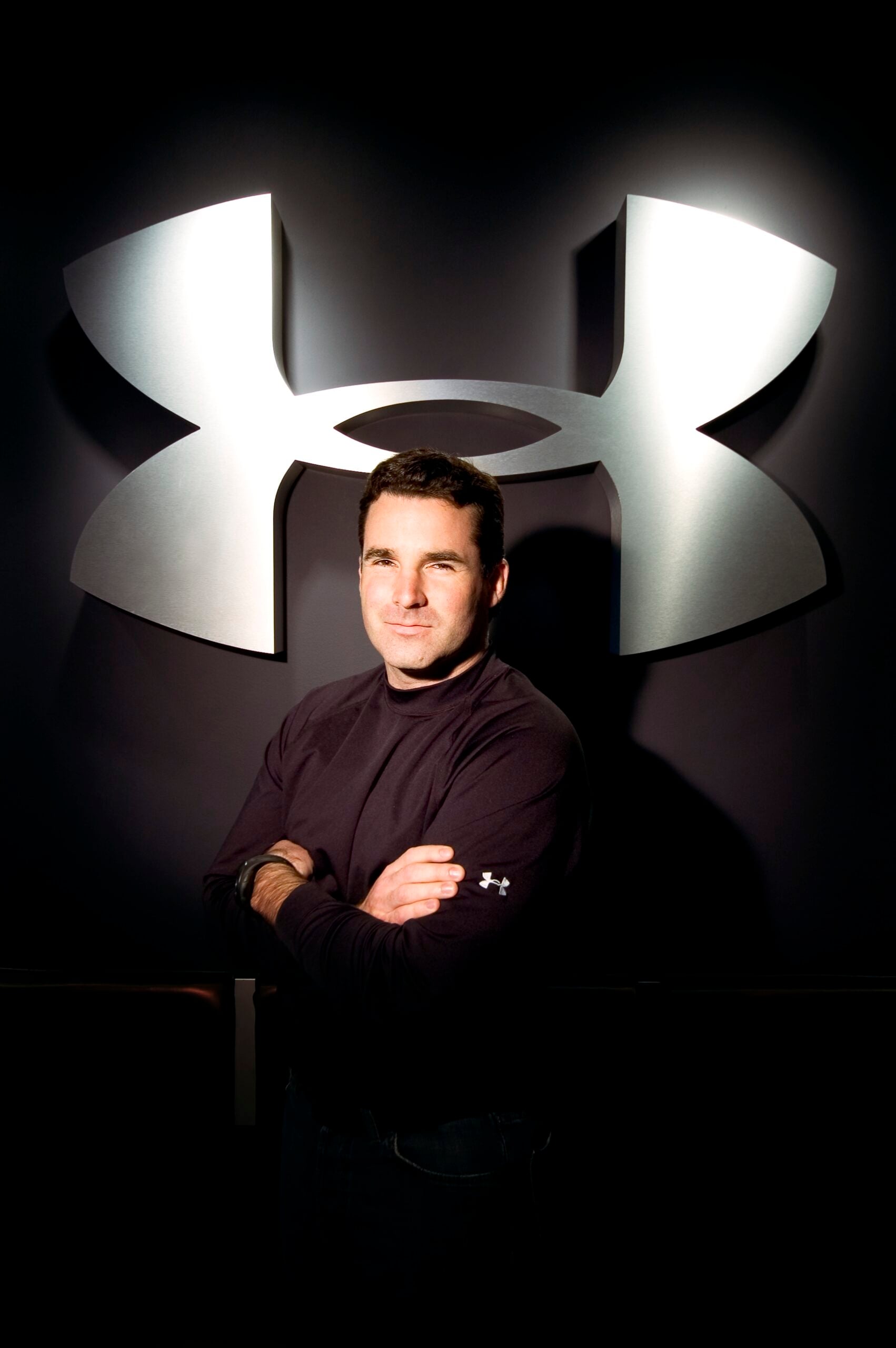 A man in a black long-sleeve shirt stands with arms crossed in front of a large, metallic Under Armour logo on a dark background.