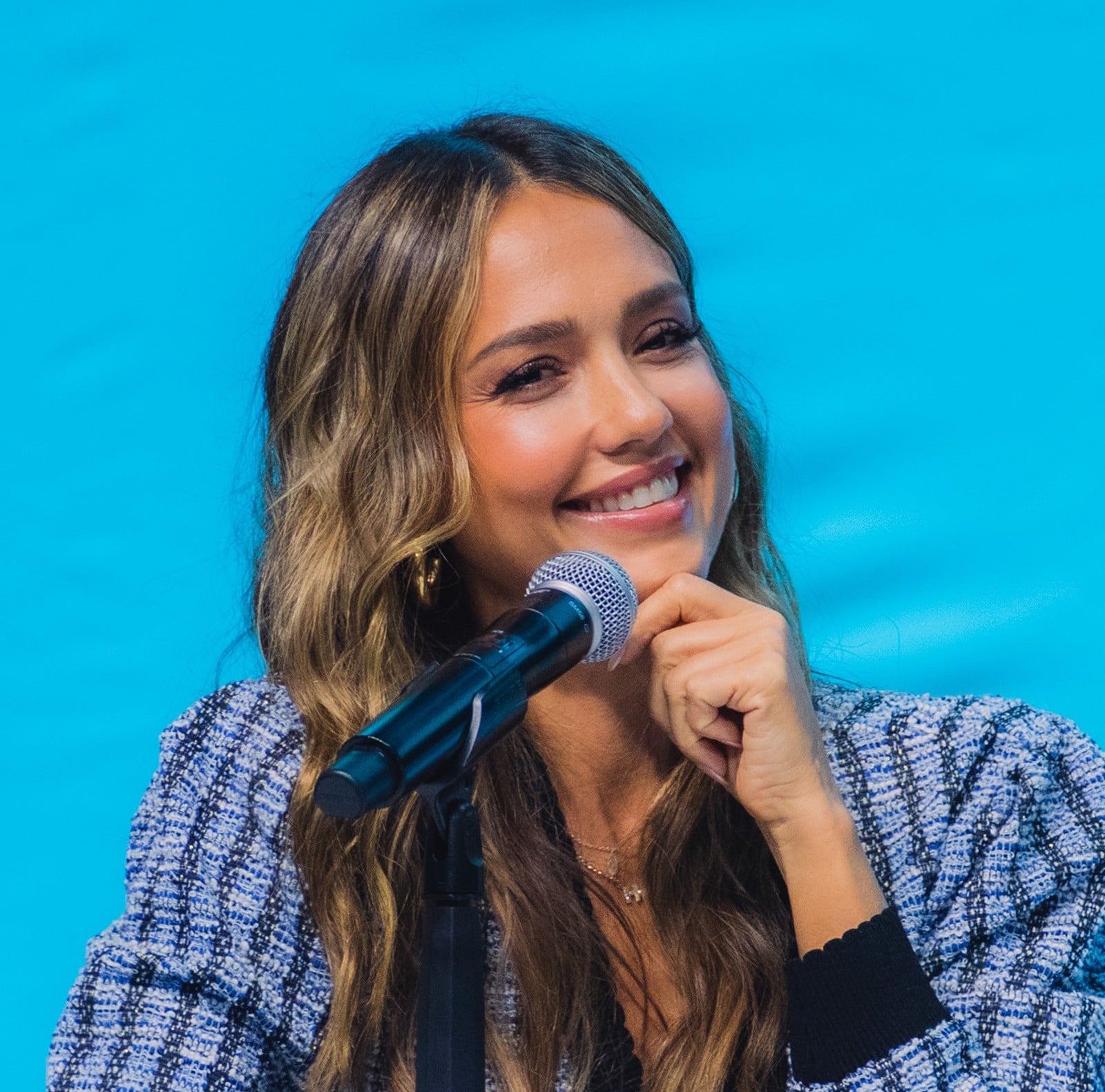 A woman with long wavy hair smiles while holding her chin, sitting in front of a microphone against a bright blue background. She wears a patterned jacket and appears to be at a speaking event or panel.