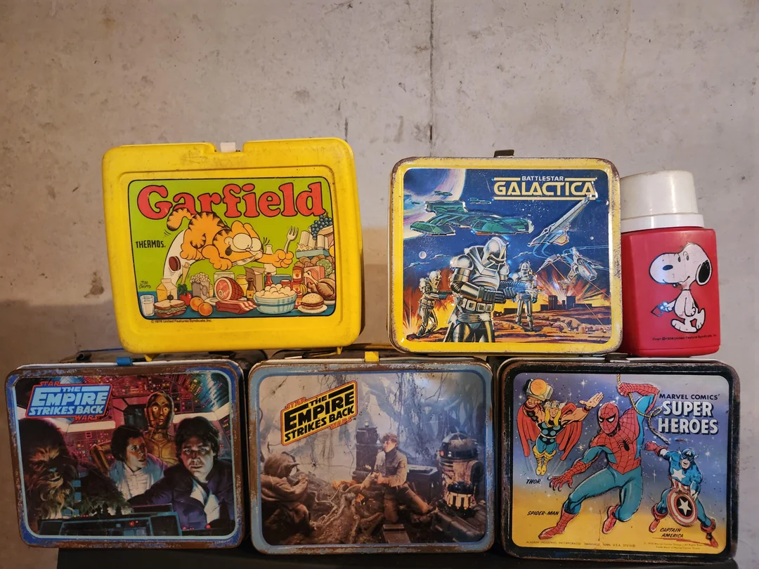 Five vintage lunchboxes featuring Garfield, Battlestar Galactica, Star Wars, and Marvel superheroes are displayed with a red Snoopy thermos against a plain background.