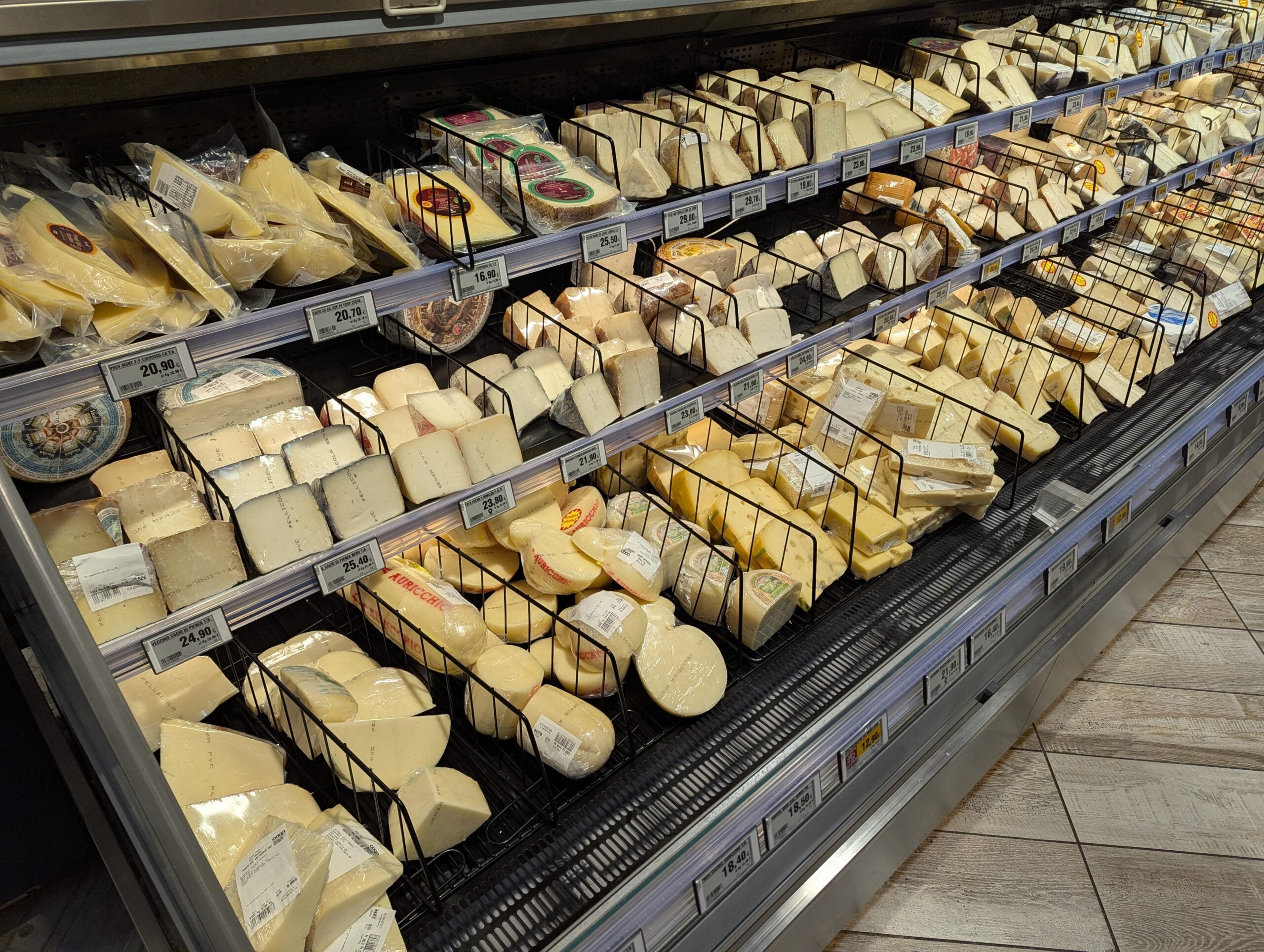 Shelves in a grocery store refrigerator display a wide variety of cheeses, each packaged in different shapes and sizes, with price tags and labels visible on the wire racks.