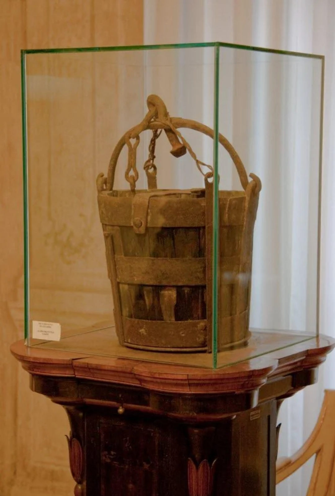 A weathered wooden bucket with metal bands and handle is displayed inside a glass case atop a dark wooden stand in a museum setting.