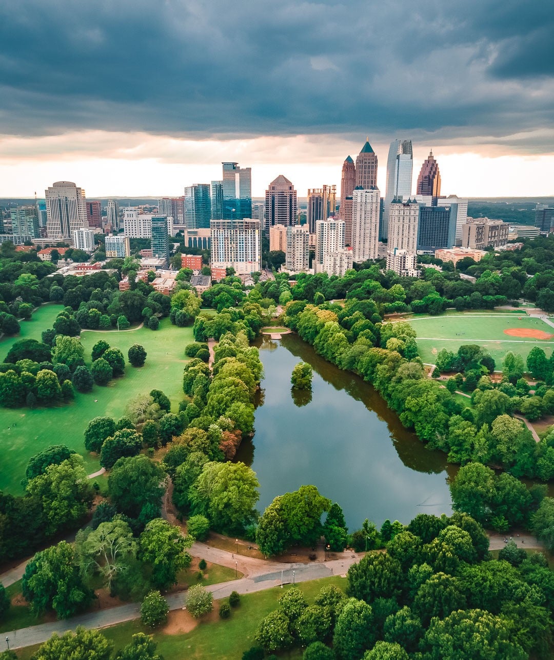 Aerial view of a city skyline with tall buildings, a large green park, a reflective pond, and cloudy skies overhead.