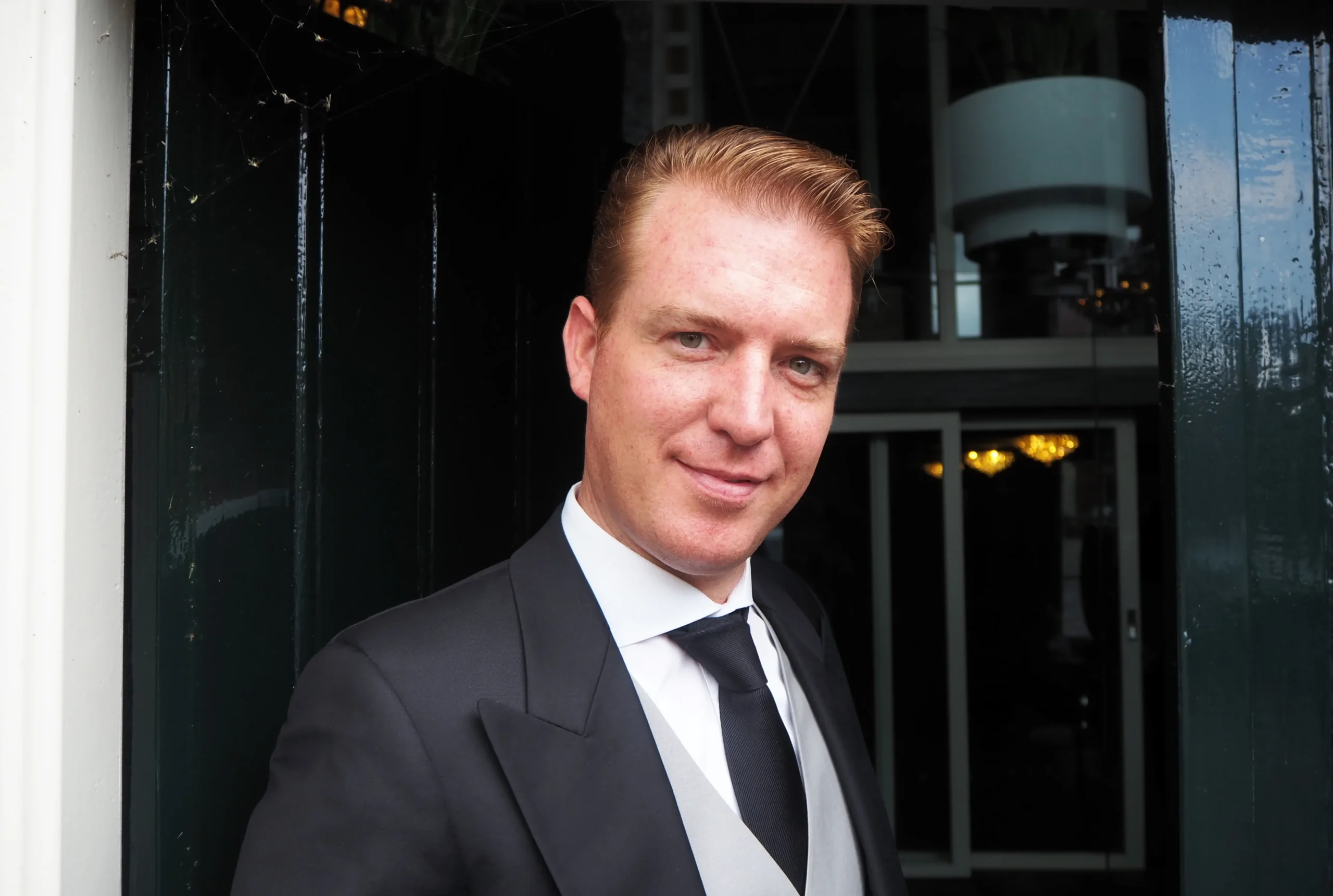 A man with short, reddish-blond hair wearing a black suit, white shirt, and black tie stands smiling in front of a dark doorway with glass panels and indoor lighting visible behind him.