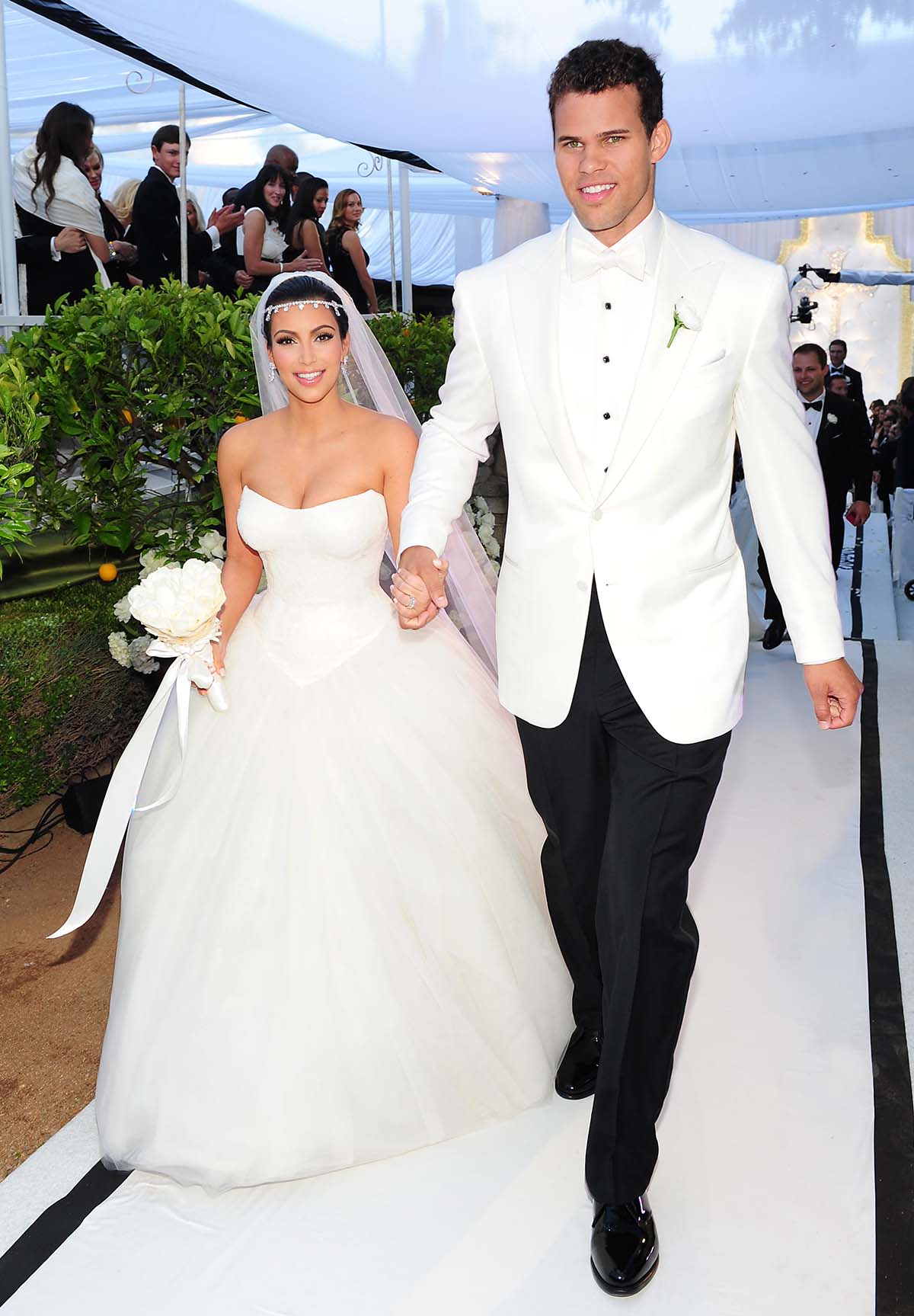 A bride in a strapless white gown and veil holds hands with a groom in a white tuxedo as they walk down an outdoor aisle surrounded by guests and greenery.