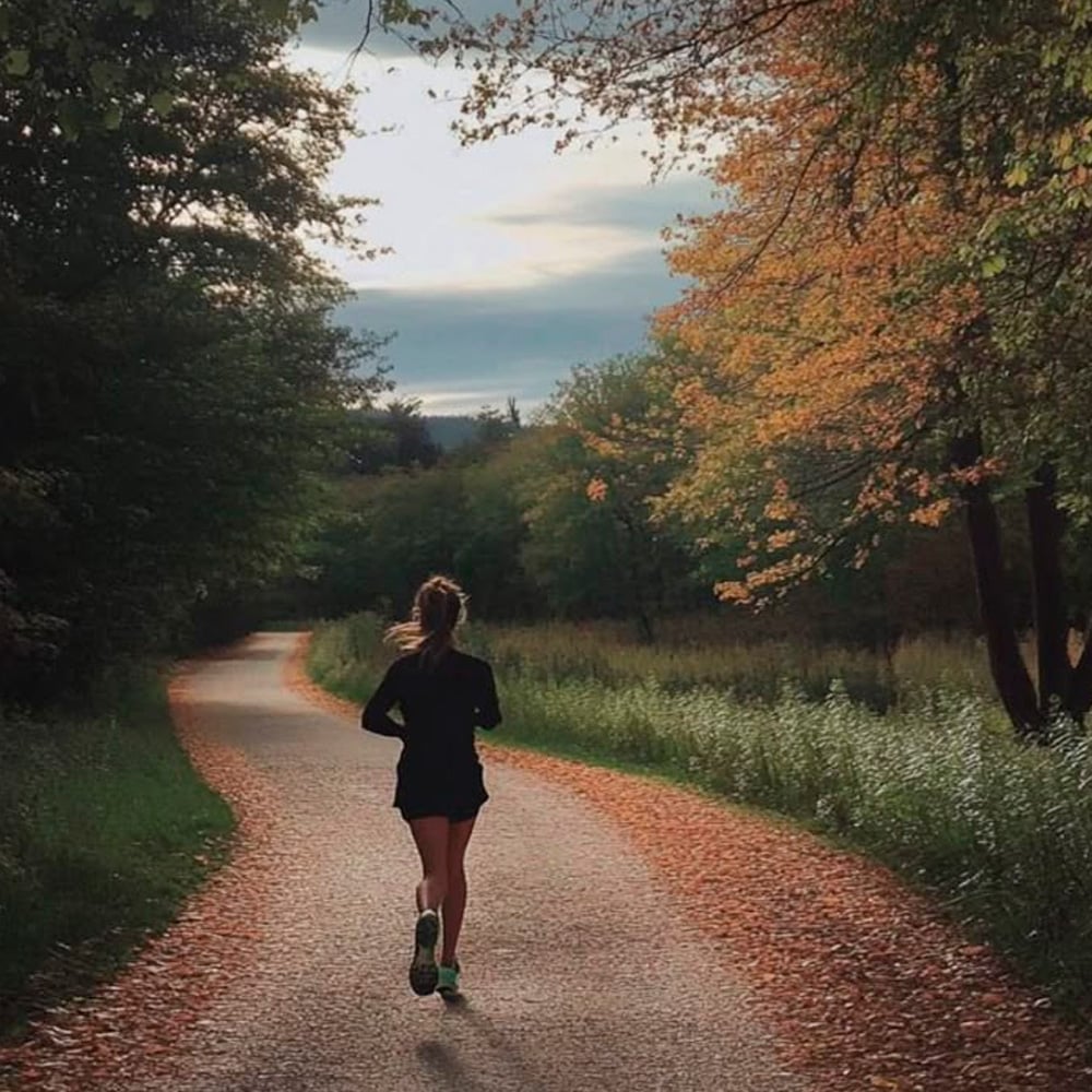 A person jogs along a winding path surrounded by trees with autumn foliage, under a cloudy sky. The path is bordered by grass and fallen leaves, creating a peaceful, scenic atmosphere.