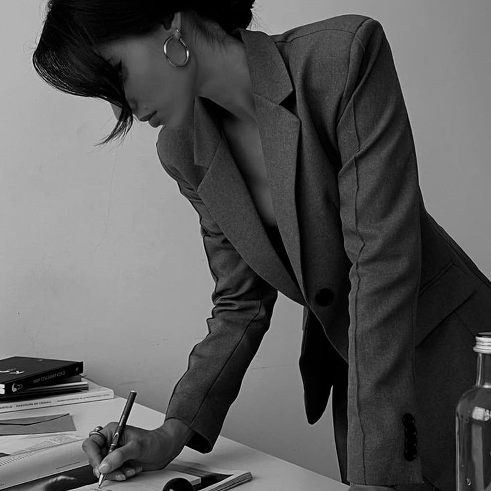 A woman in a blazer leans over a desk, writing in a notebook. Her hair partly covers her face, and she wears hoop earrings. On the desk are books, a bottle, and papers. The photo is in black and white.