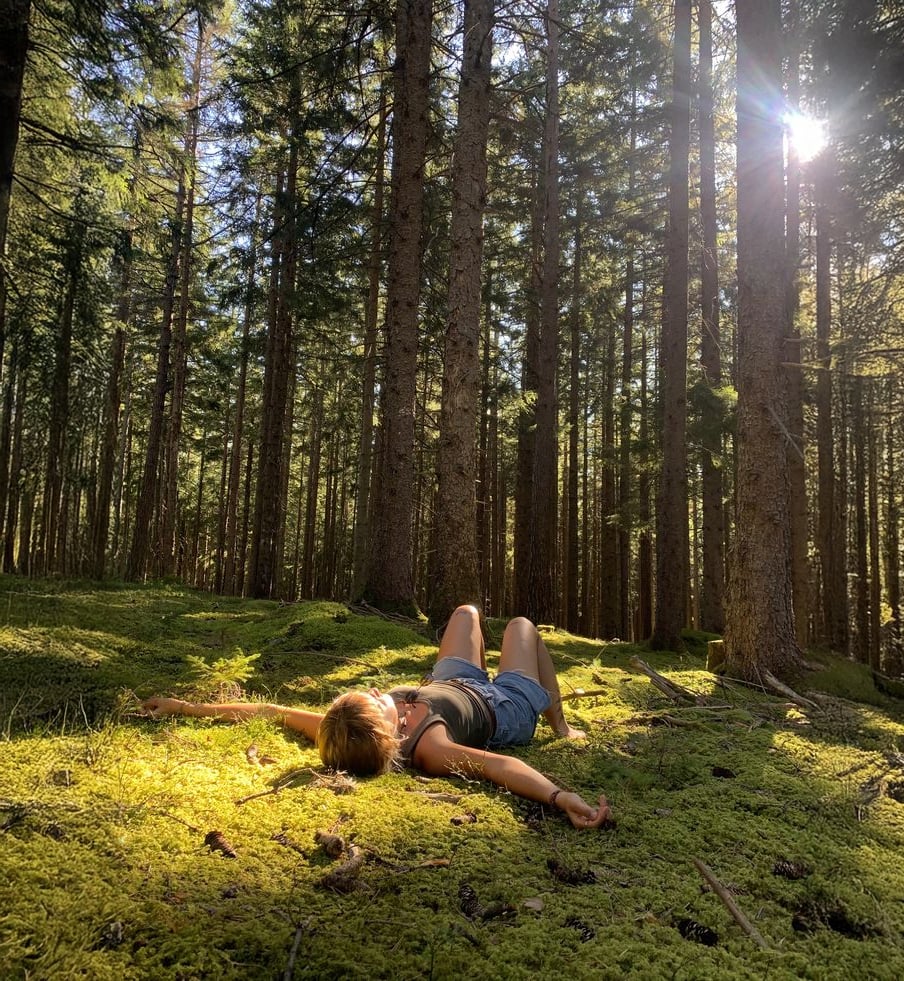 A person lies on their back on a sunlit mossy forest floor, surrounded by tall trees with sunlight filtering through the branches.