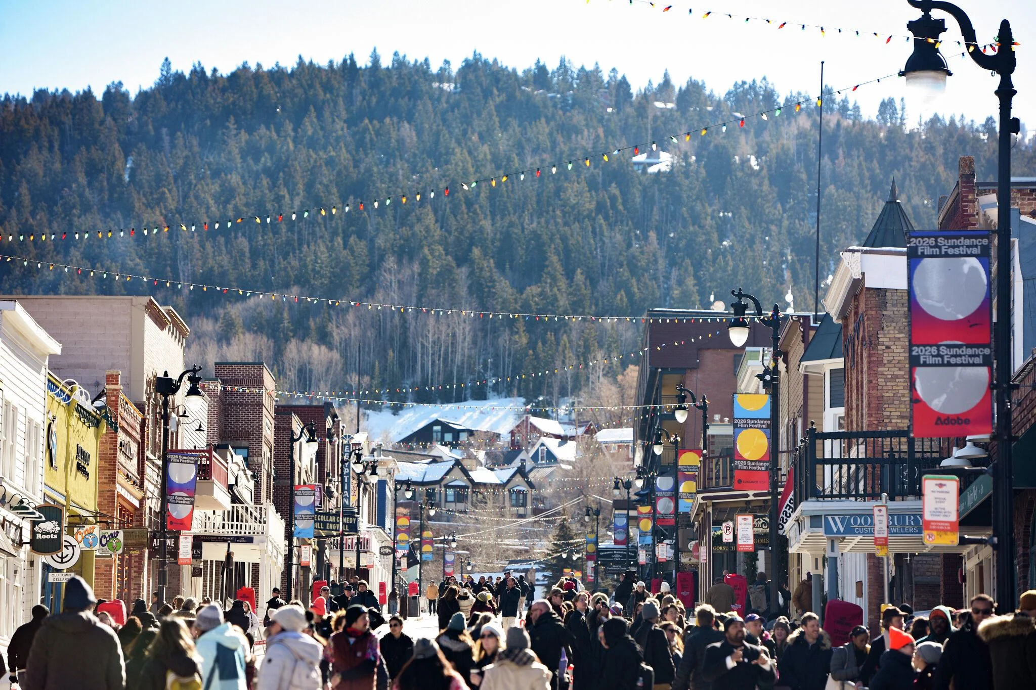A crowded street in a mountain town, lined with shops and colorful banners, with snow-covered trees and hills in the background. People in winter clothing walk under string lights on a sunny day.