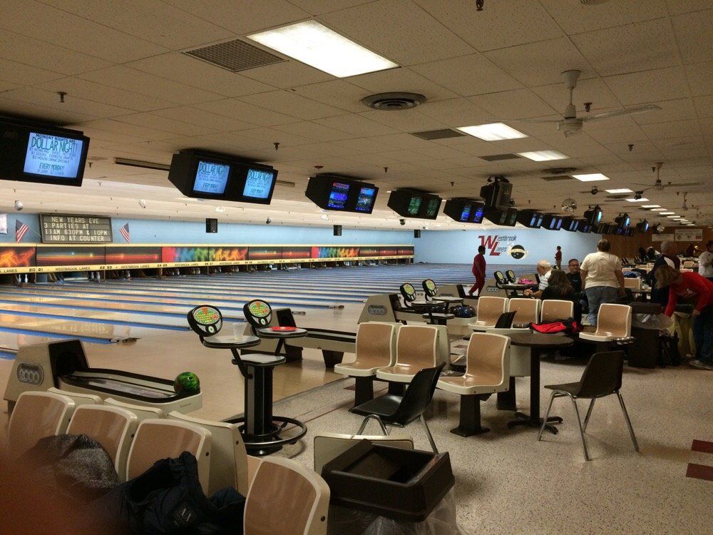A bowling alley with several empty lanes and scoring monitors overhead, tan chairs and tables in the foreground, and a group of people gathered near the lanes in the background.