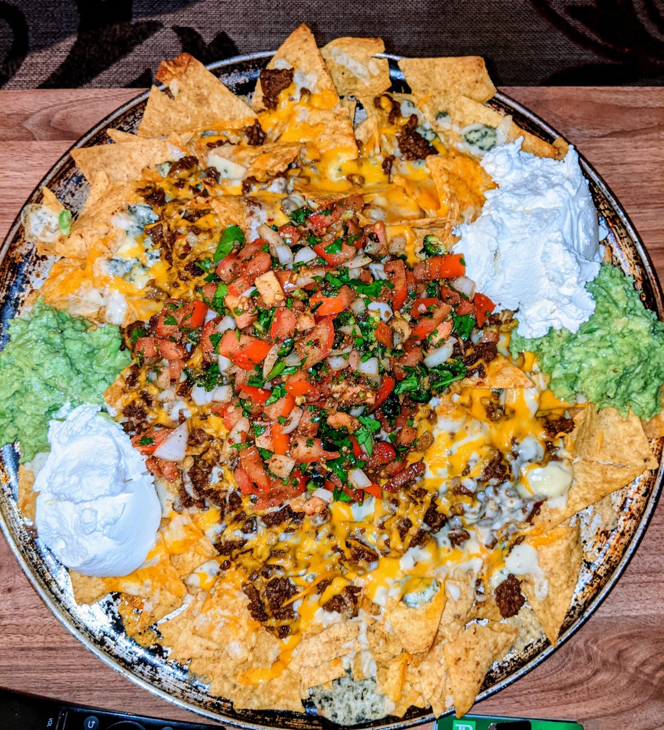 A large plate of loaded nachos topped with melted cheese, seasoned ground beef, pico de gallo, guacamole, and sour cream on a wooden table.
