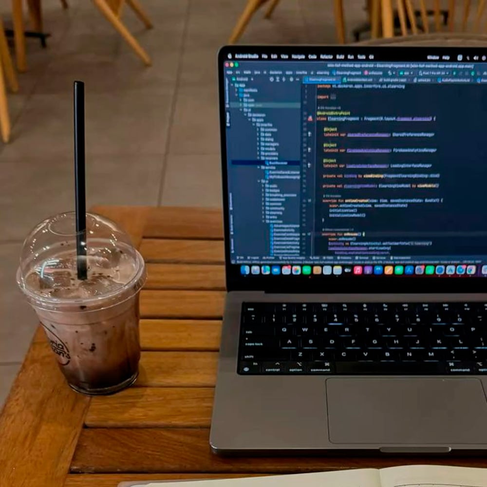 A laptop displaying code sits on a wooden table next to a plastic cup of iced chocolate with a straw. Part of a notebook is also visible at the bottom of the image. Wooden chairs are seen in the background.
