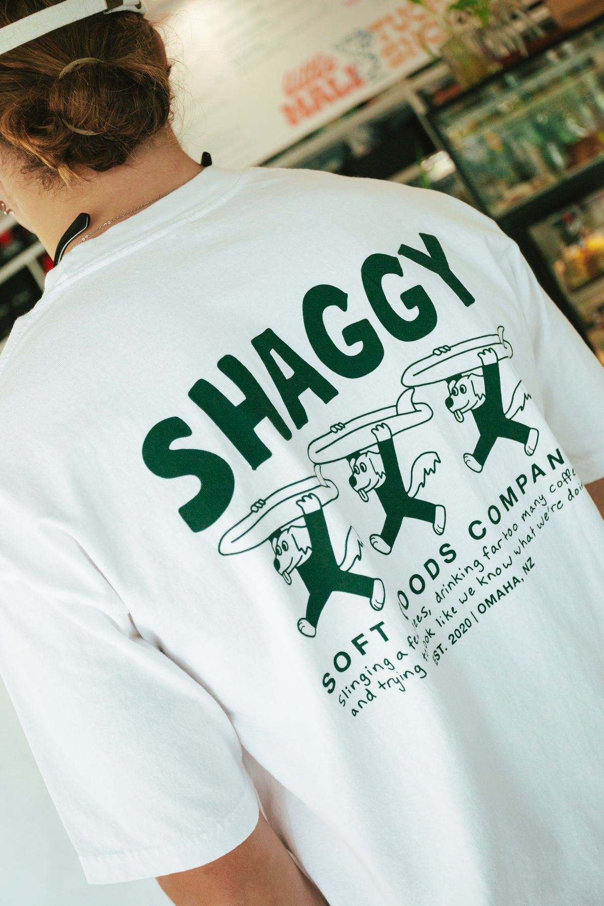 A person wearing a white T-shirt with "SHAGGY" in large green letters and cartoon figures holding pizza slices, along with text promoting Soft Foods Company and its mission, is shown from behind.