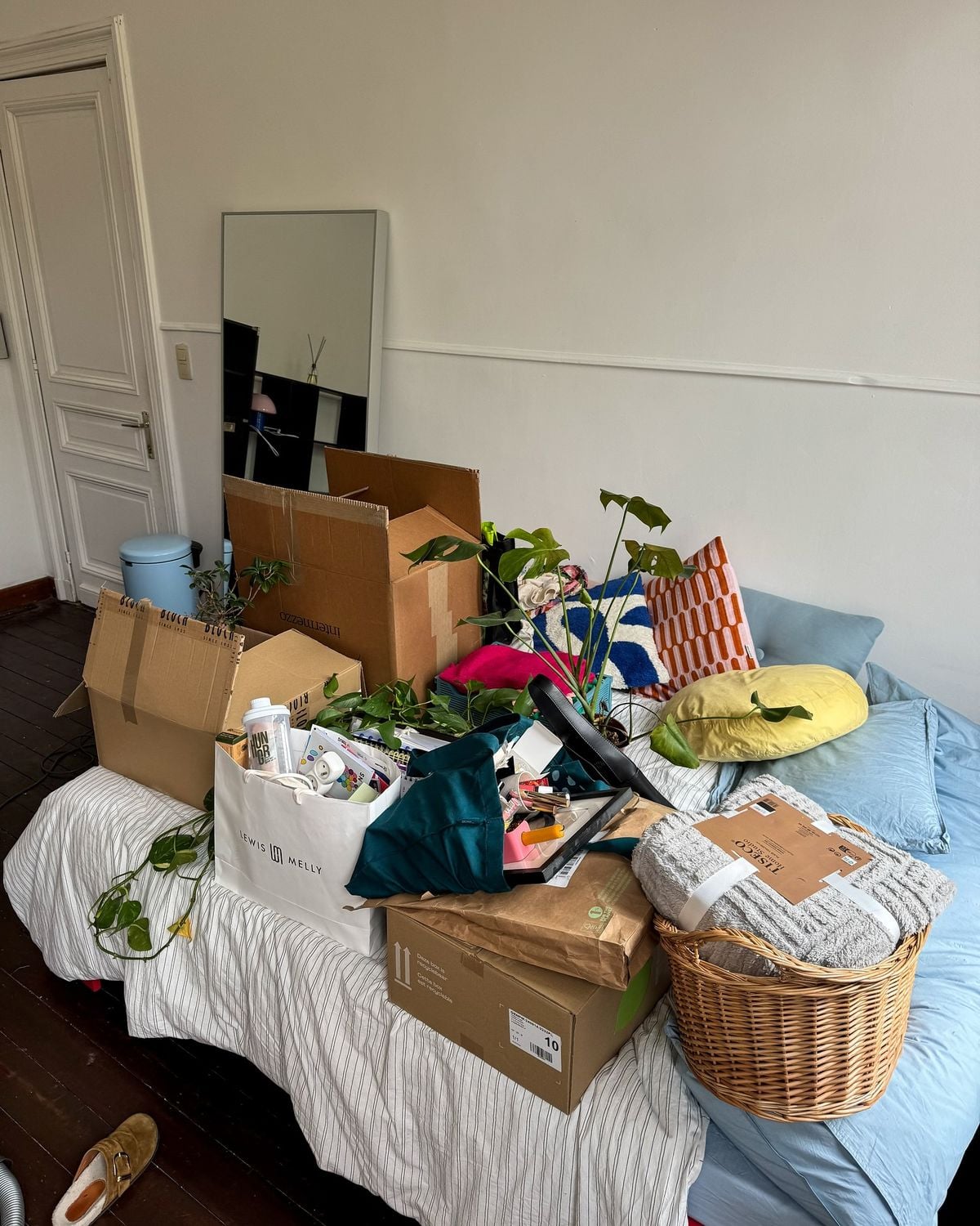 A messy bed covered with boxes, bags, plants, and household items. A mirror leans against the wall, and a pillow, blanket, and decor pieces are visible. The room has wooden floors and white walls.
