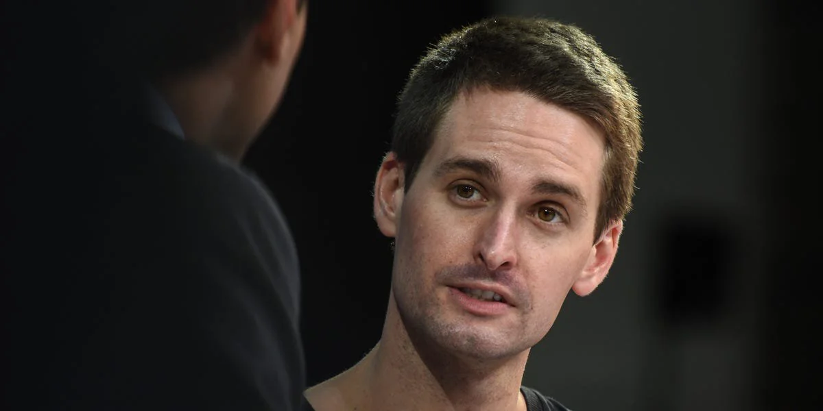 A man with short brown hair wearing a black shirt is speaking to another person, who is partially visible and blurred in the foreground. The background is dark and out of focus.