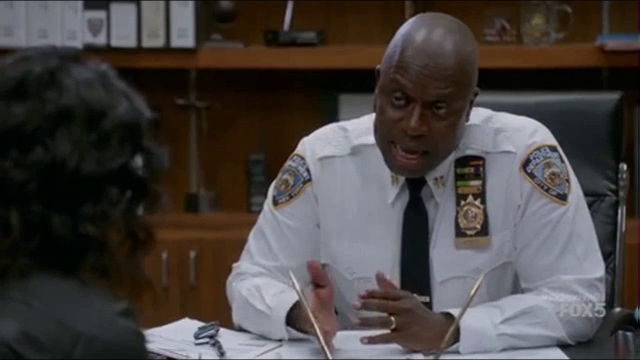 A police captain in uniform sits at a desk in an office, talking seriously to another person whose back is to the camera. Papers and folders are visible on the desk.
