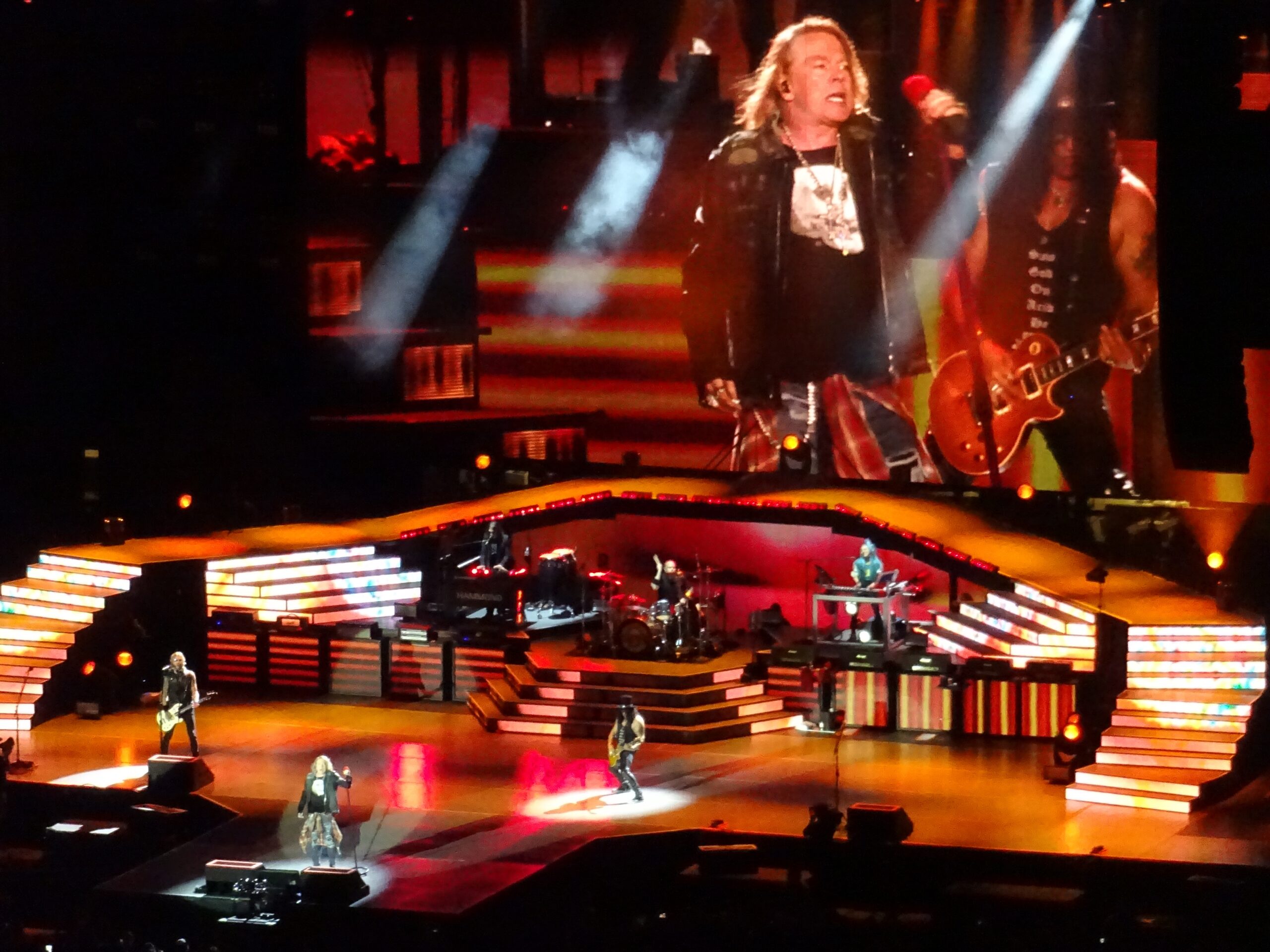 A rock band performs on a brightly lit stage with colorful lights and a large screen displaying the lead singer and guitarist. The audience watches as musicians play various instruments amid dynamic stage effects.