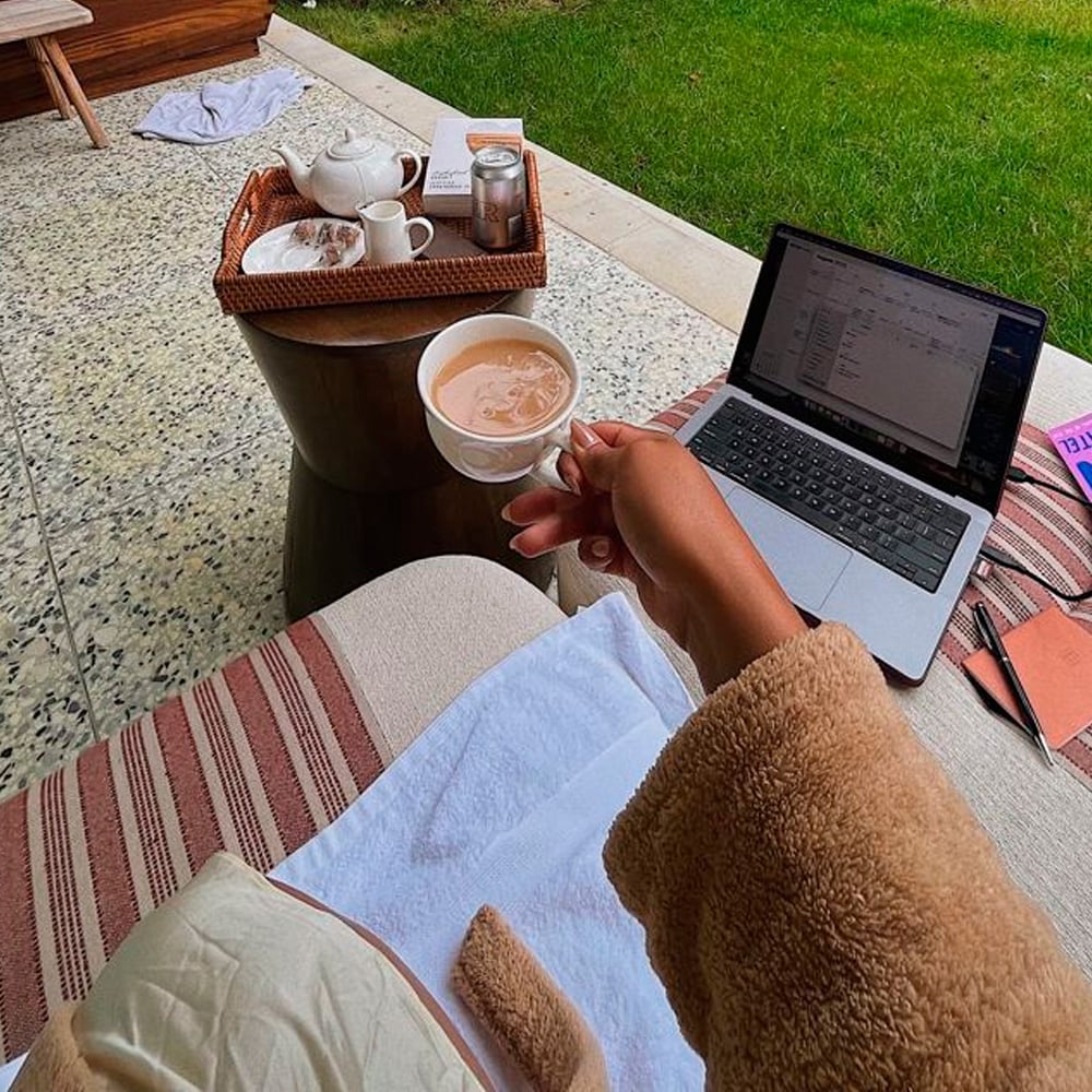 A person in a beige robe holds a cup of coffee next to a laptop on a small outdoor table, with a tray of tea and a green lawn in the background.