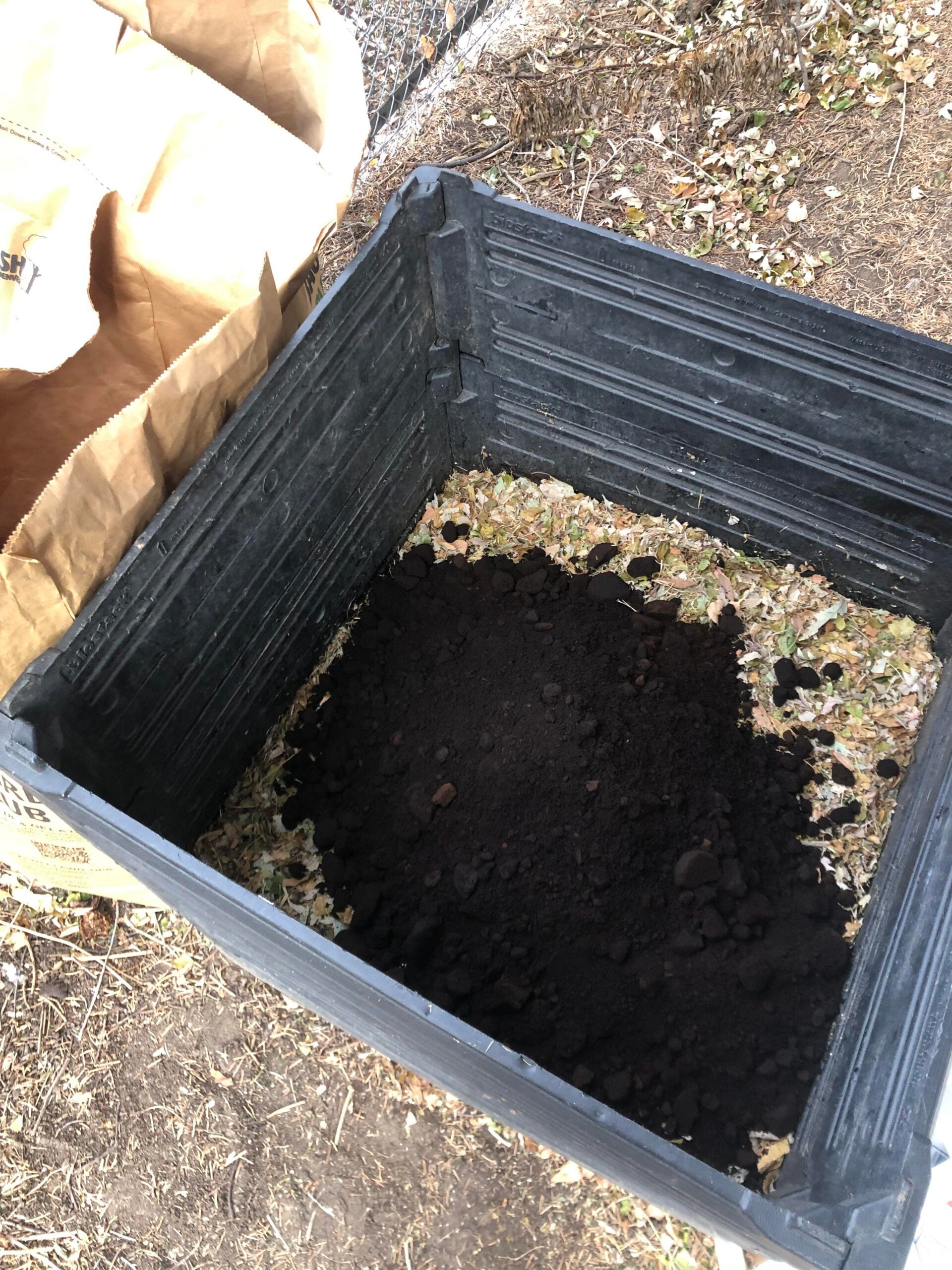 A black plastic compost bin contains a layer of dried leaves with a pile of dark compost or soil in the center. A brown paper bag is visible next to the bin, and dry grass surrounds the area.