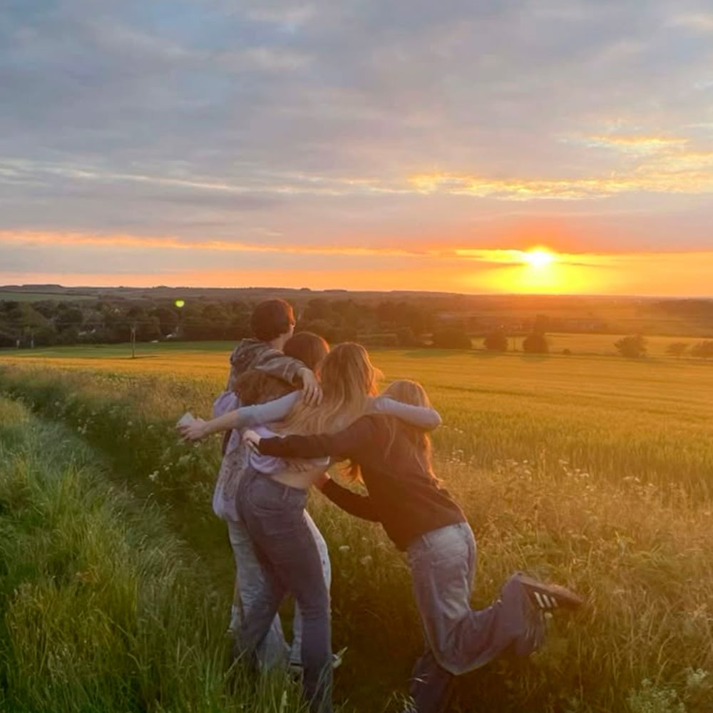 Four people stand together in a grassy field at sunset, embracing each other and looking toward the horizon. The sky is partly cloudy, and the landscape is bathed in warm, golden light.