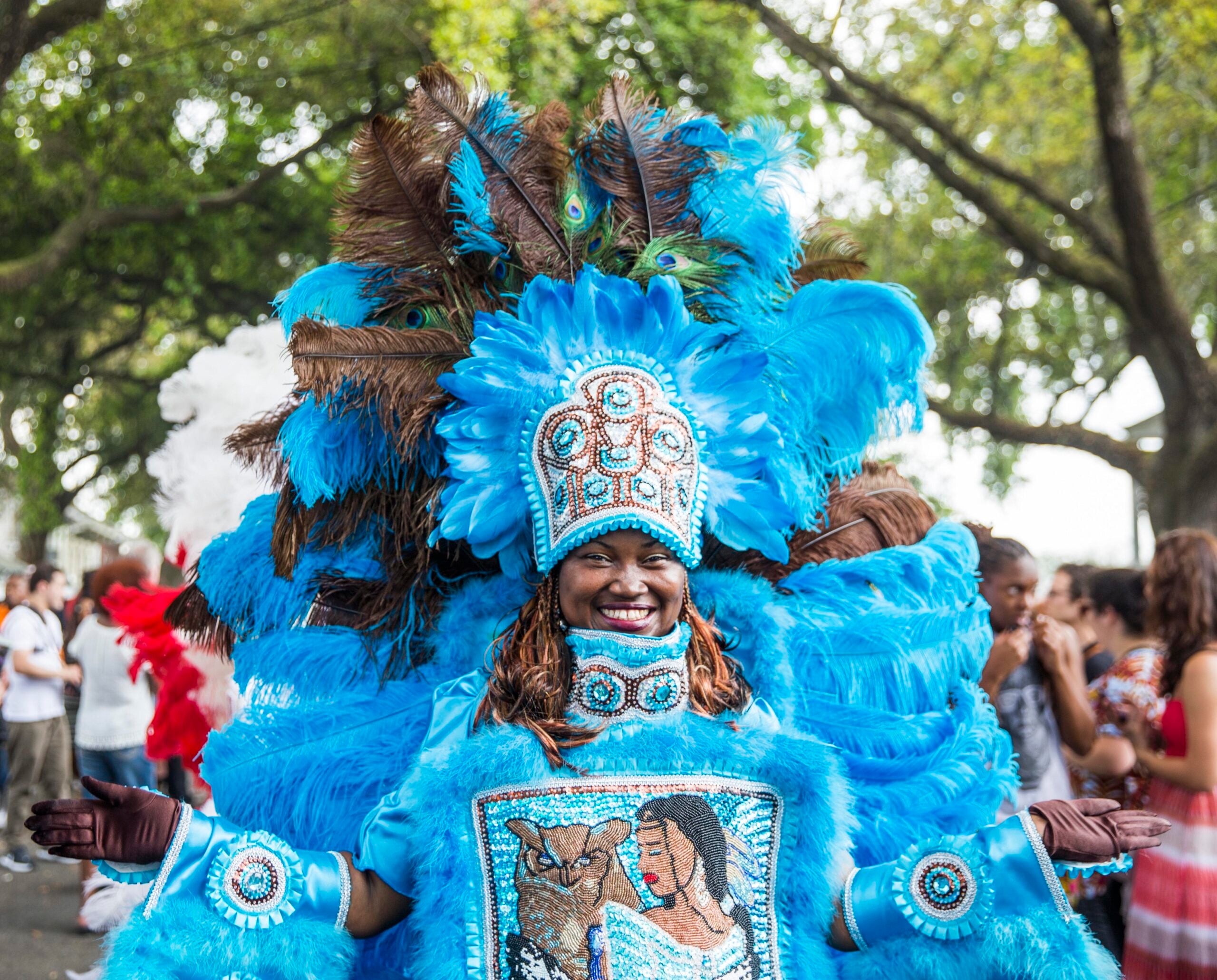 A person smiles widely while wearing an elaborate bright blue feathered costume with beadwork and a matching headdress, standing outdoors at a lively event with people and trees in the background.