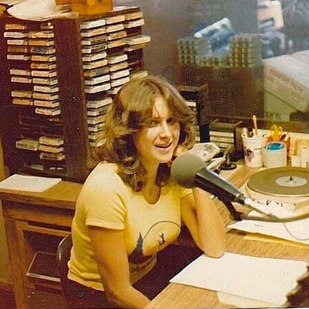 A young woman with wavy hair sits at a desk in a radio studio, speaking into a microphone. She is wearing a yellow T-shirt. In the background are audio cassette tapes, papers, and studio equipment.