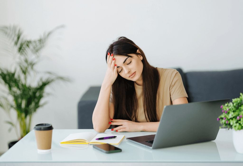 A woman sits at a desk with her eyes closed and her hand on her forehead, looking tired. She is surrounded by a laptop, notebook, smartphone, and a coffee cup, with a plant in the background.