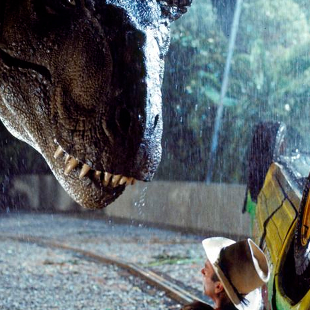 A large dinosaur bares its teeth at a person wearing a hat, who is sitting on the ground beside an overturned vehicle in the rain, surrounded by trees.