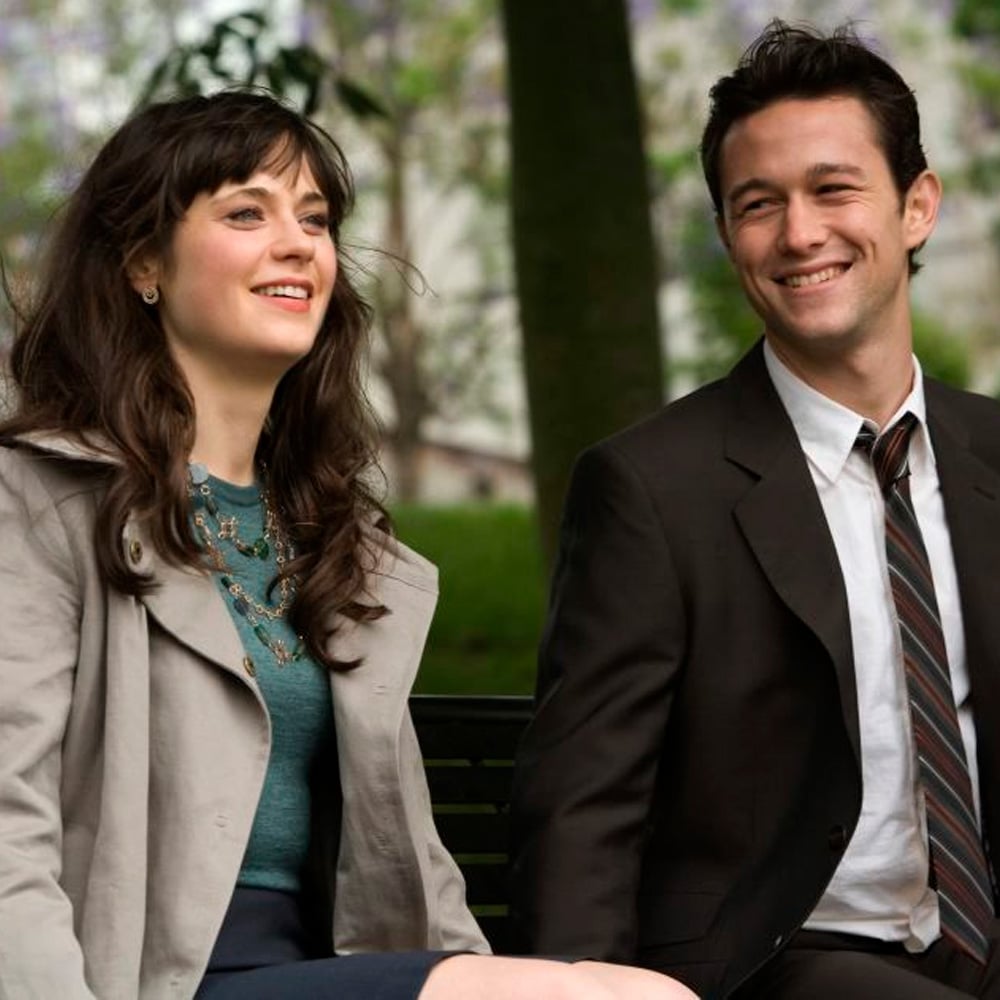 A woman and a man sit on a bench outdoors, smiling. The woman has long brown hair and wears a light coat over a teal top, while the man wears a dark suit and striped tie. Trees and blurred buildings are in the background.