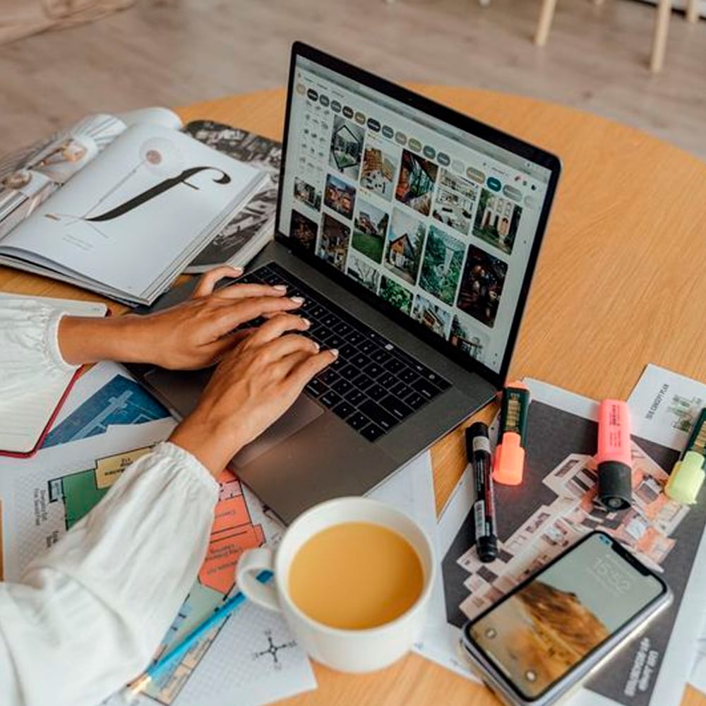 A person types on a laptop at a cluttered table with magazines, papers, highlighters, a smartphone, a pen, and a cup of coffee. The laptop screen displays a photo gallery.