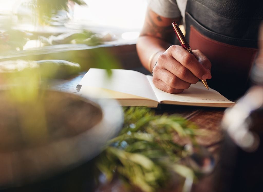 A person is writing in a notebook with a pen at a wooden table. The scene includes blurred green plants in the foreground and soft natural light coming from the left.