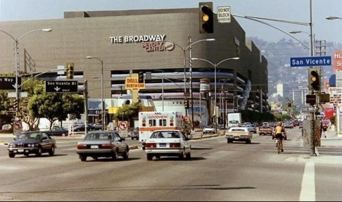 Cars and a cyclist move through a busy city intersection near The Broadway Beverly Center, with street signs for San Vicente Blvd and Beverly Blvd visible, and hills in the background.