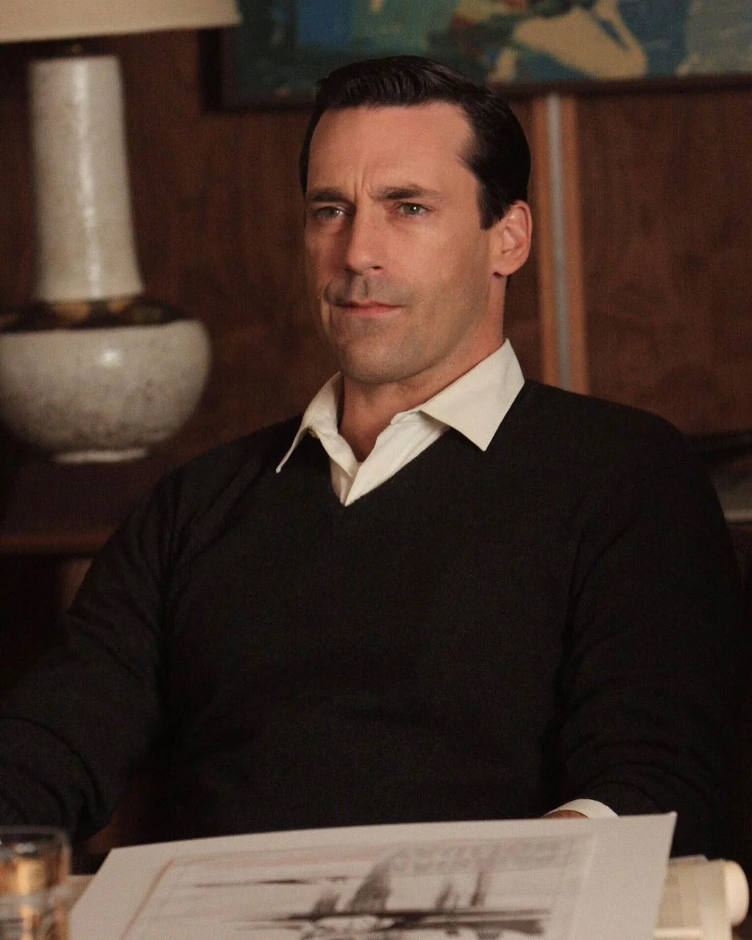 A man with dark hair, wearing a white shirt and dark sweater, sits at a desk with papers in front of him in a dimly lit office setting. A lamp and wood-paneled wall are in the background.