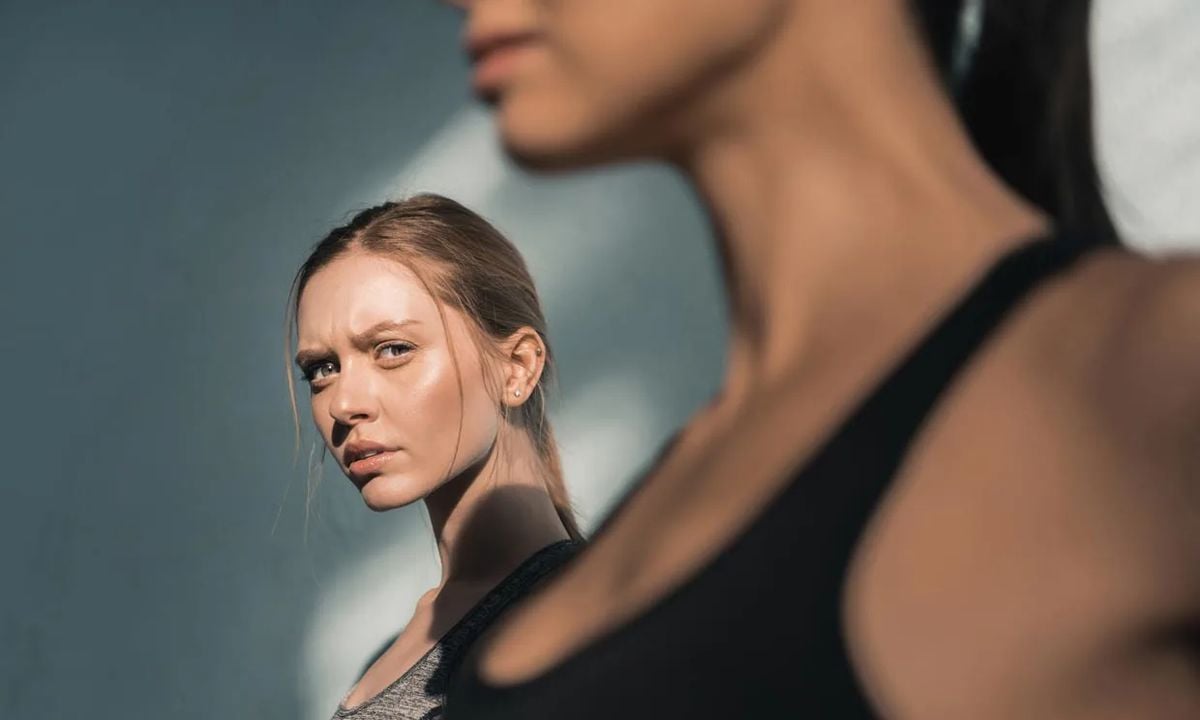 A woman with light hair gazes intently at another woman in the foreground, who is blurred. Both wear athletic tops and stand against a neutral background with soft lighting and shadows.