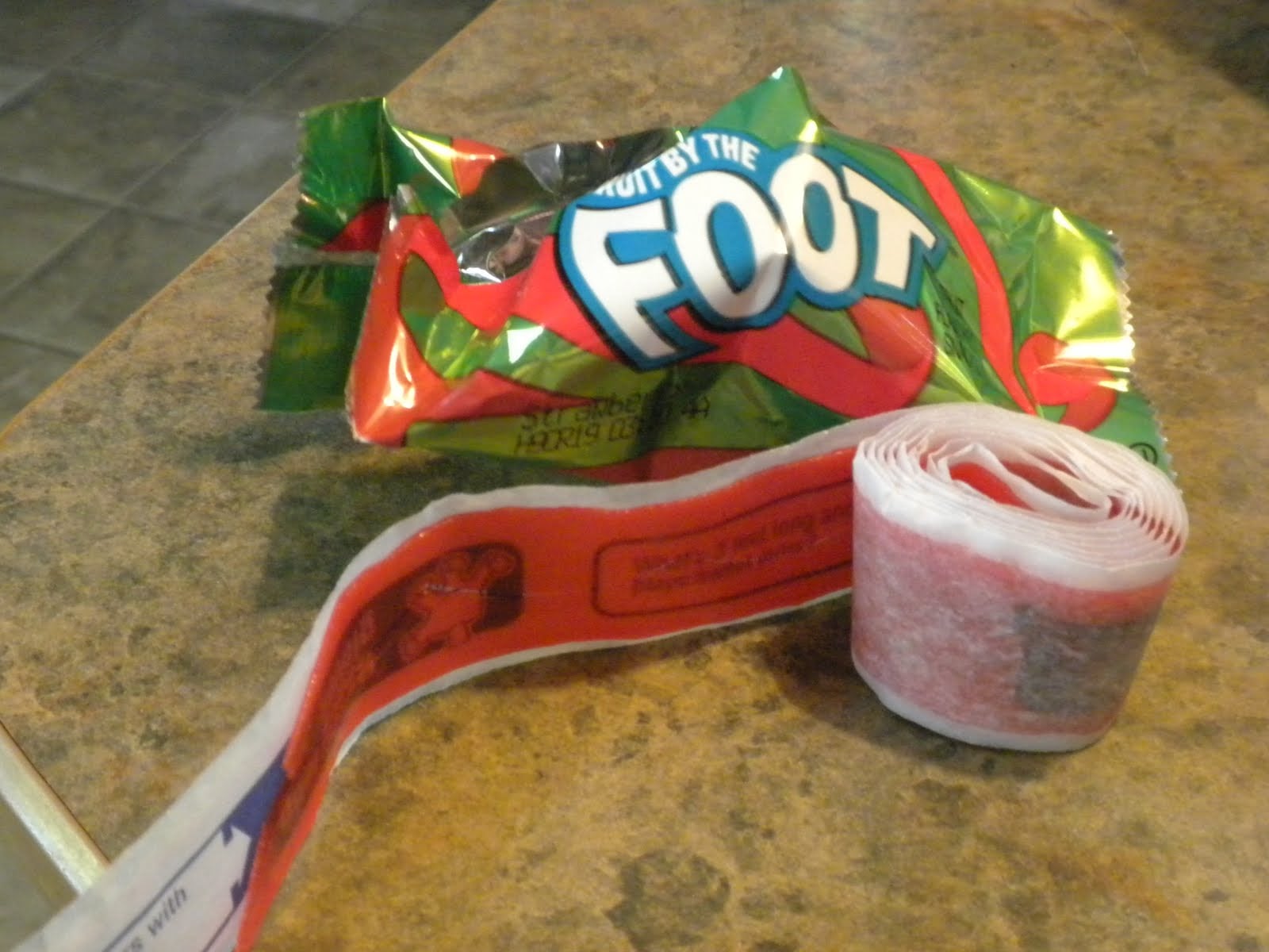 A rolled-up strip of red Fruit by the Foot candy sits on a countertop next to its colorful, partially opened green wrapper.
