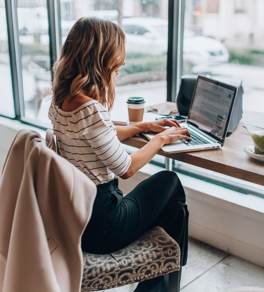 A woman sits at a café table by a large window, working on a laptop. She holds a coffee cup and has a beige coat draped over her chair. Sunlight streams in, and there are cars visible outside.