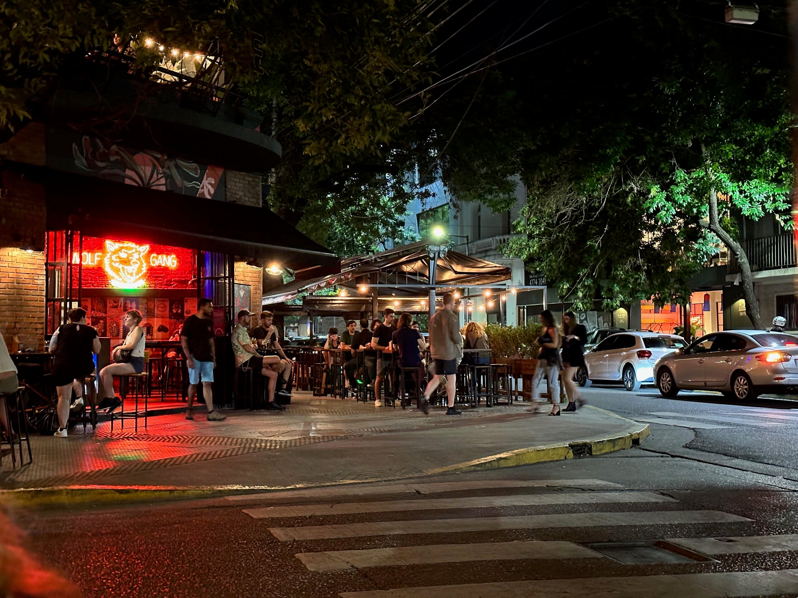 People sit and stand outside a lively bar with a red neon sign at night. Trees and string lights decorate the scene, and cars drive by on the street, creating a vibrant urban atmosphere.
