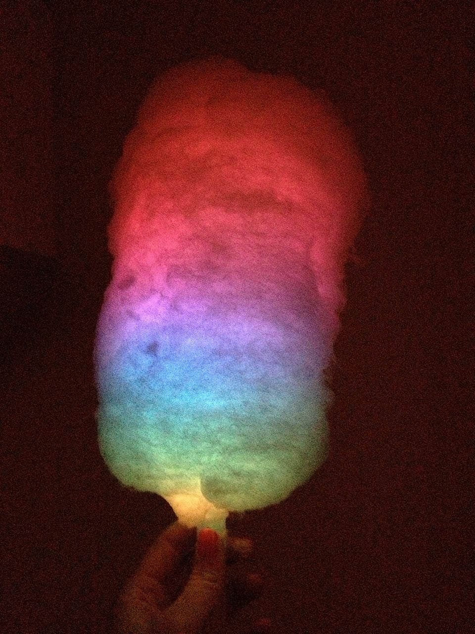 A hand holds a stick of cotton candy that glows in vibrant colors, including red, pink, purple, blue, and green, against a dark background.