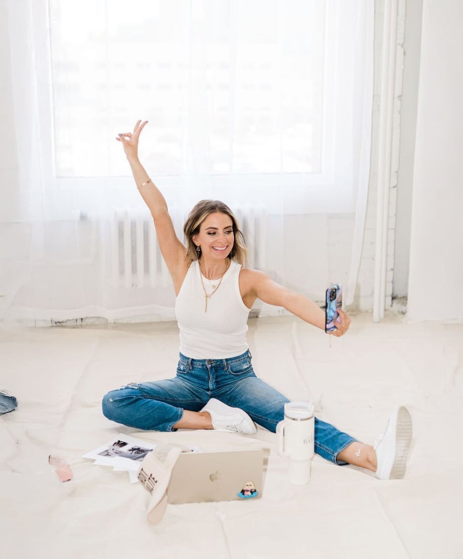 A woman in a white tank top and jeans sits cross-legged on the floor, smiling and holding up her phone for a selfie. She is surrounded by a laptop, books, a water bottle, and sunglasses in a bright, airy room.