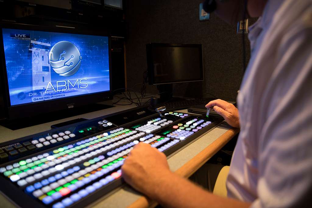 A person operates a control panel with many buttons and sliders in a broadcast studio. A monitor displays a live video feed titled “ARMS Dr. [Name]” with a futuristic graphic and blue background.