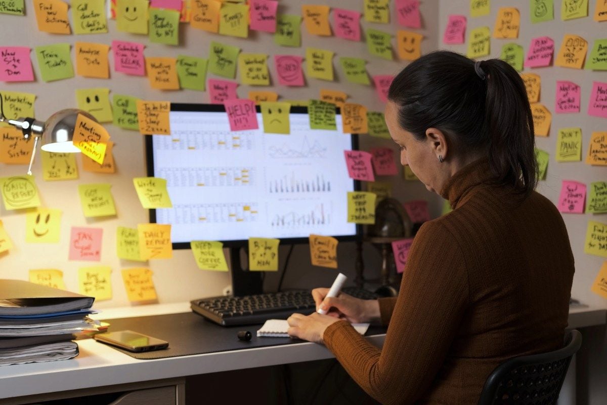 A woman sits at a desk covered in papers and a phone, writing in a notebook. The wall and computer monitor in front of her are covered with colorful sticky notes and charts, indicating a busy, organized workspace.