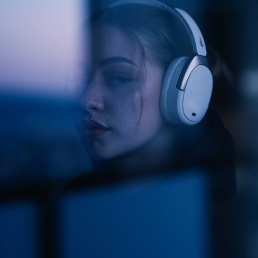 A young woman with long hair wears large white headphones, gazing thoughtfully through a window with soft blue and purple light reflecting on her face. The image is slightly blurred and moody.