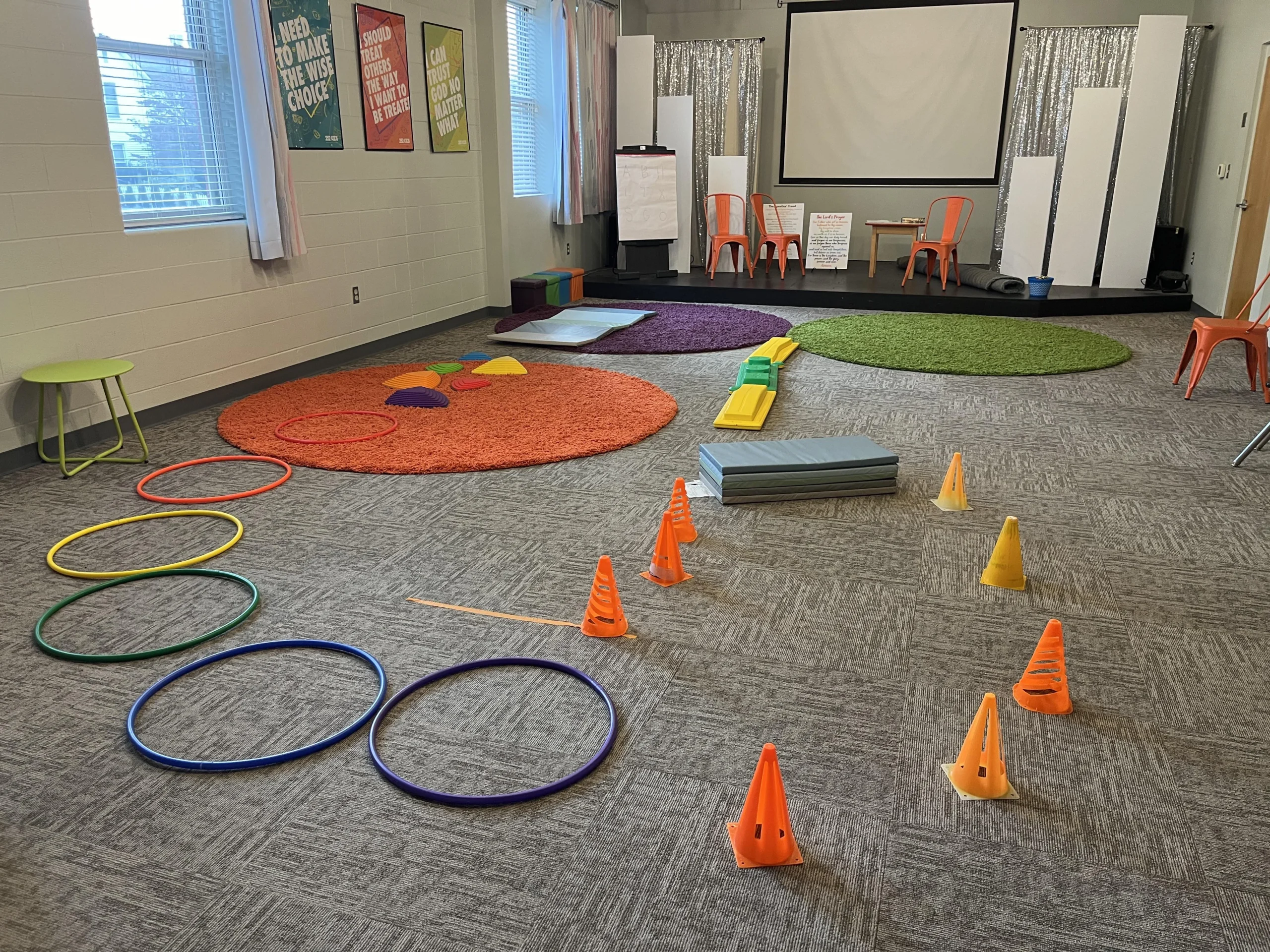 A classroom with colorful rugs, hoops, cones, balance beams, and mats arranged as an obstacle course. Orange chairs line the back, and motivational posters hang on the walls. A large screen is at the front.
