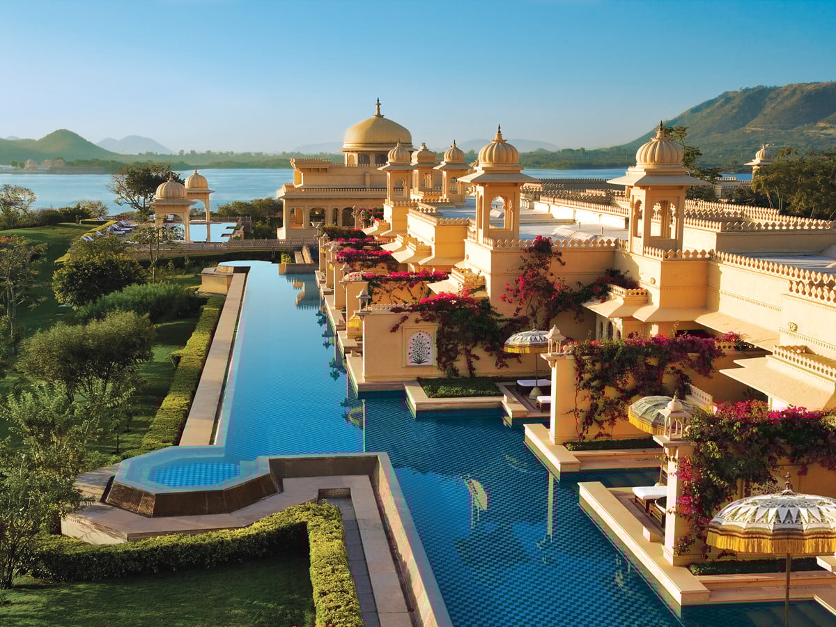 A luxurious palace-style hotel with domes and arches, surrounded by lush greenery and reflected in a long, blue pool, set beside a serene lake and mountains under a clear blue sky.