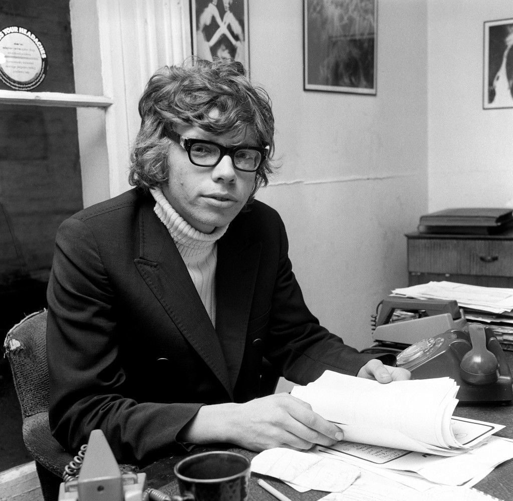 A young man in glasses and a suit jacket sits at a cluttered desk, holding papers. He looks toward the camera. The office has scattered documents, a phone, a cup, and framed pictures on the walls.