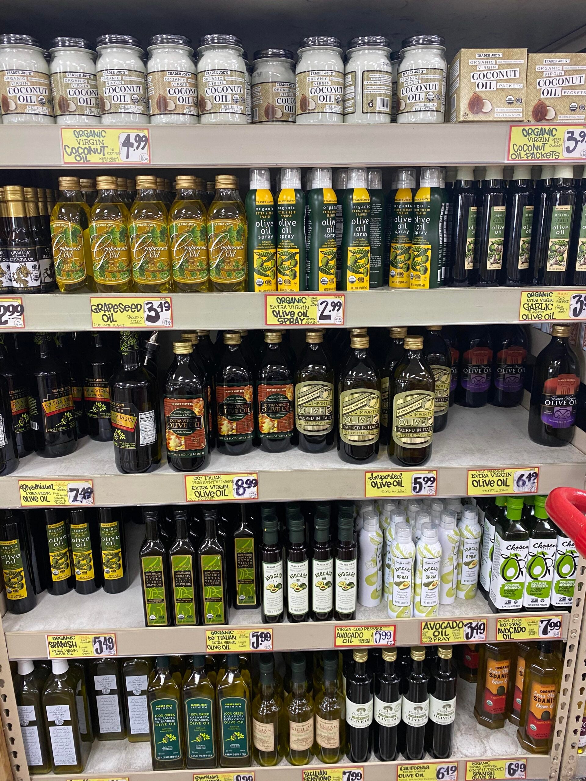 Various bottles and jars of cooking oils, including coconut, grapeseed, olive, and avocado oil, are neatly arranged on grocery store shelves with price tags displayed below each product.